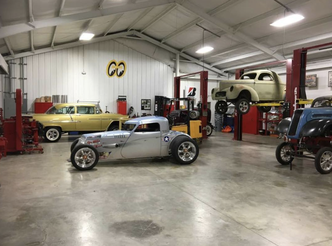 A garage with several vintage and custom cars, some on lifts and some on the floor, with a Chevrolet logo on the back wall and Ohio State emblem on the silver car.
