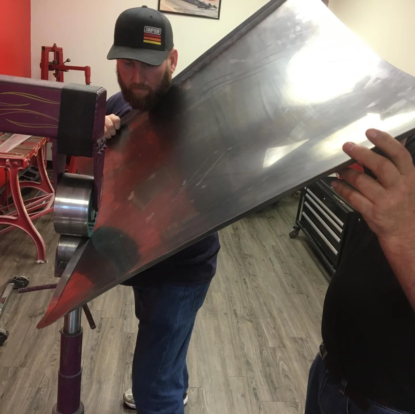 A man with a beard, wearing a black cap, is holding a large, shiny metal object, possibly a car hood or panel, in a workshop or garage. The man appears to be inspecting or working on the metal piece.