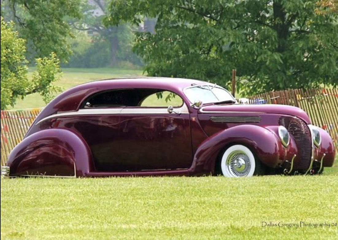 A vintage purple car with a sleek, rounded design parked on green grass in front of trees and a wooden fence.