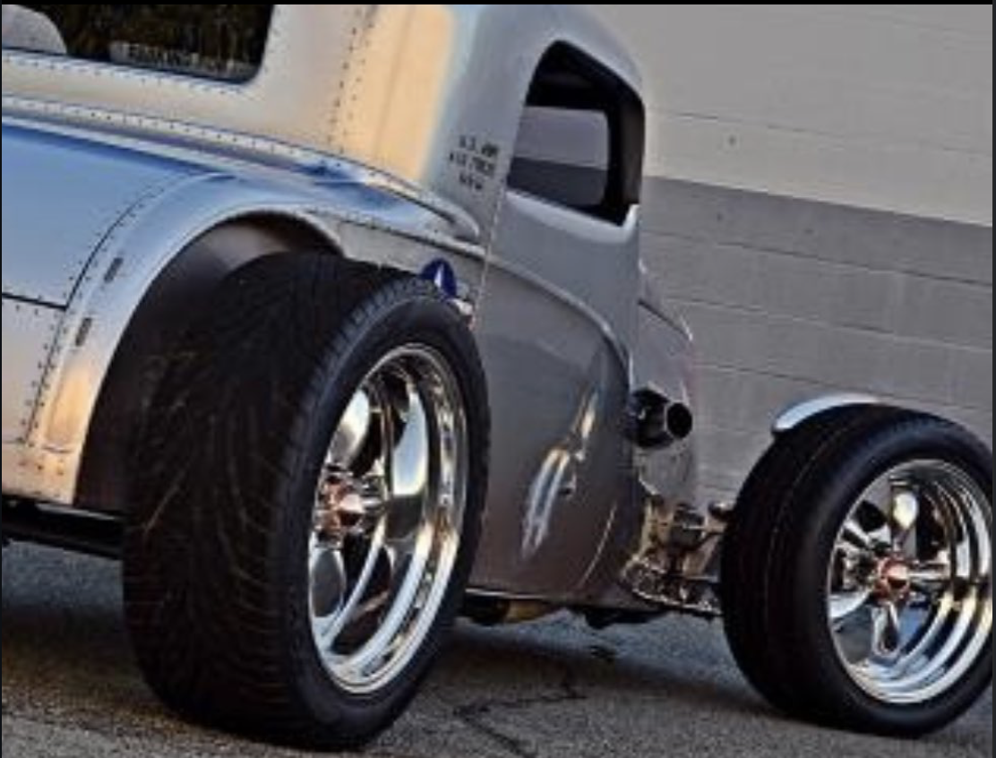 Close-up of a vintage race car with wide tires and a sleek, gray body parked near a gray wall.