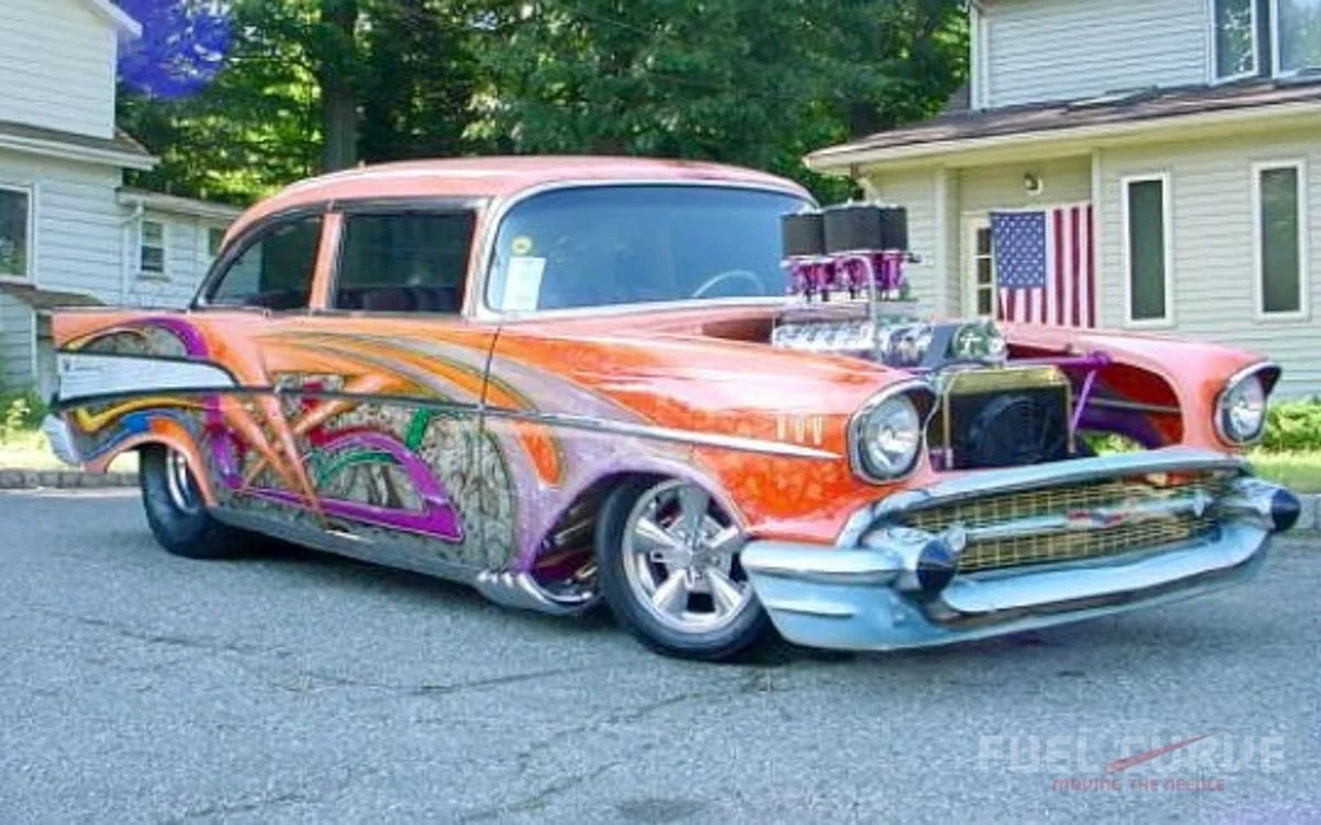 Custom vintage car with bright orange paint and purple flame design parked outside a house, featuring a large exposed engine with twin black air intakes.