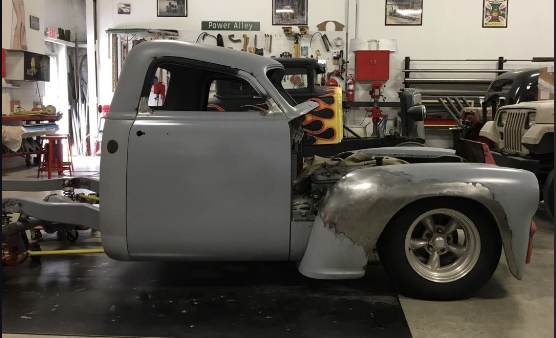 A gray vintage pickup truck body being restored in a garage workshop, with tools and other vehicle parts visible in the background.