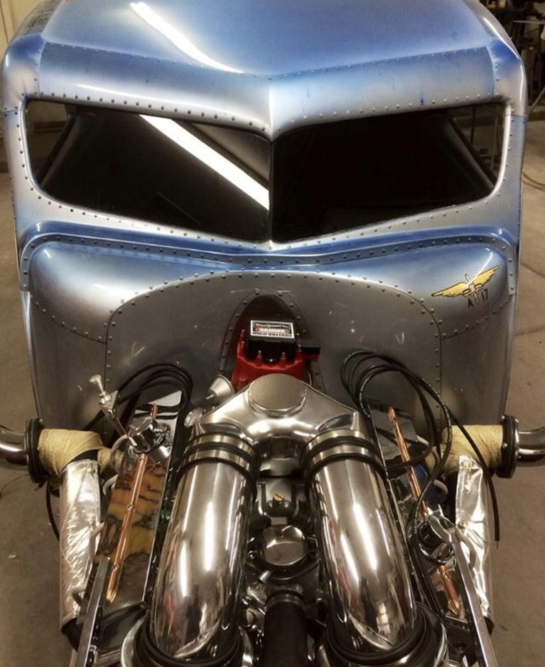 Front view of a vintage race car, featuring a metallic blue body, blacked-out windshield, and dual chrome exhaust pipes.