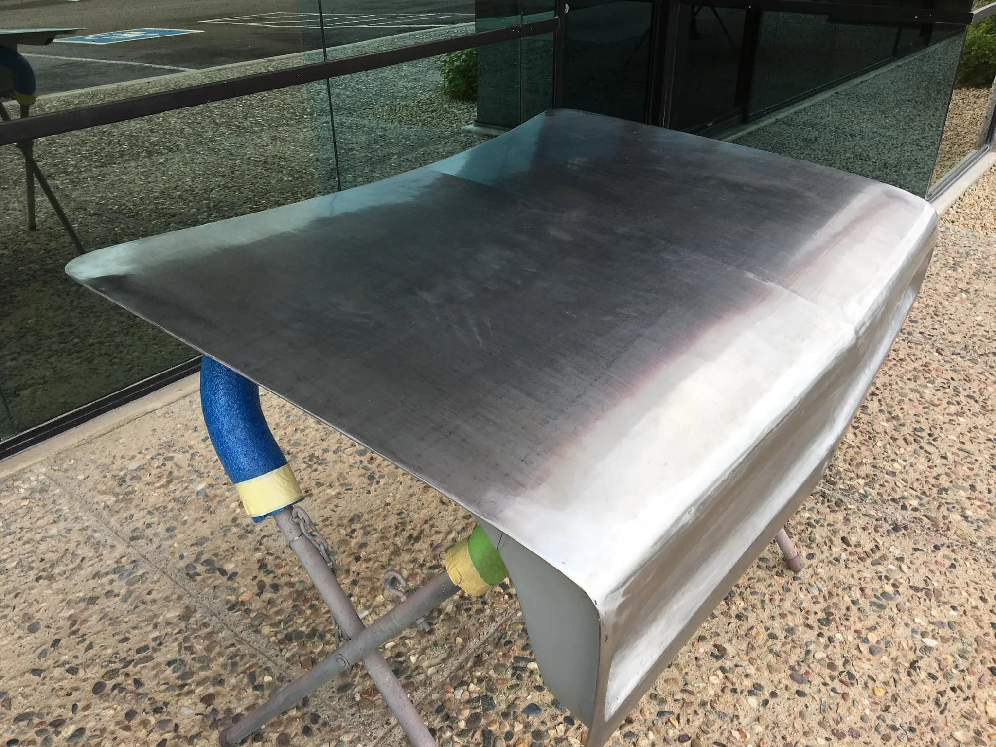A metal table with a curved edge, placed on a concrete surface outside a building with glass windows.