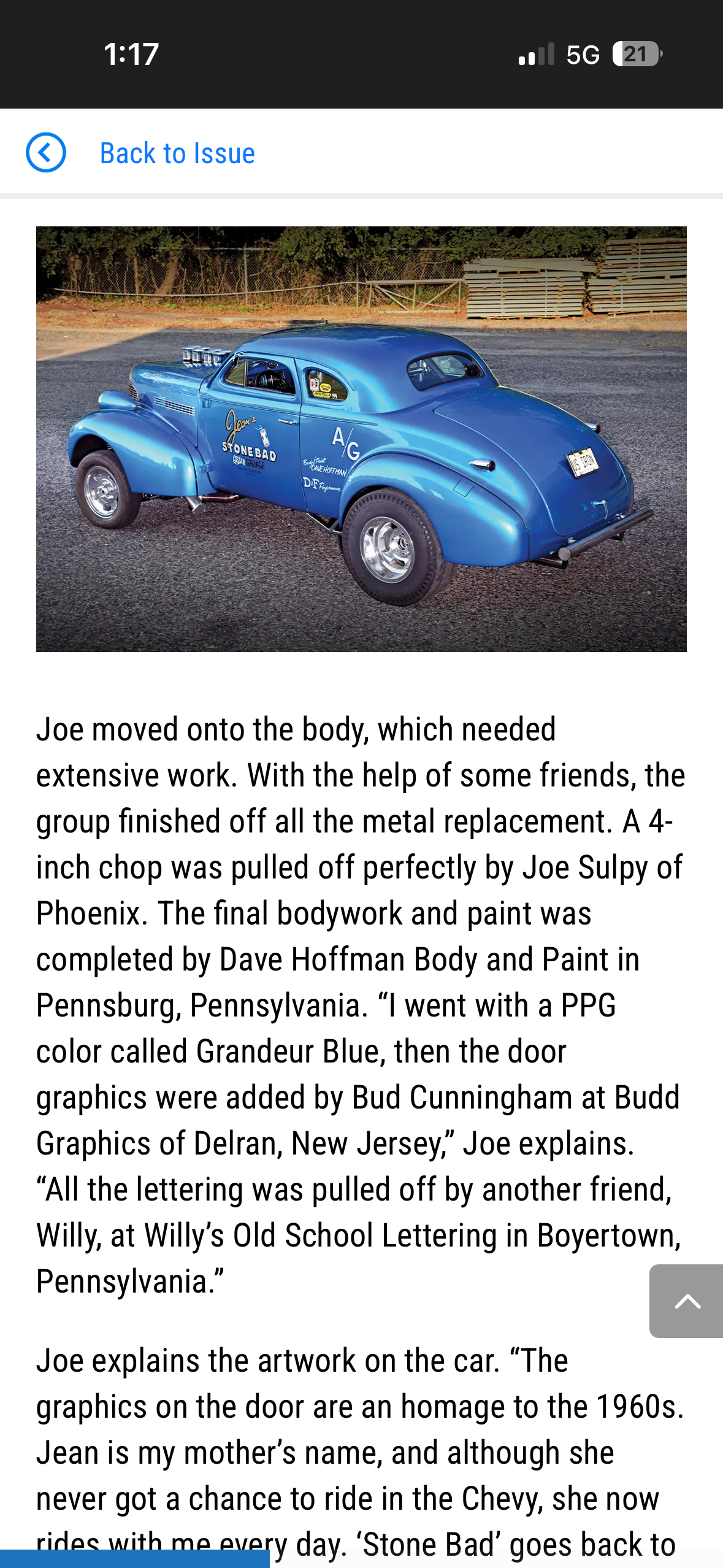 A vintage blue race car with artistic graphics and lettering, parked on a gravel surface with trees and a fence in the background.