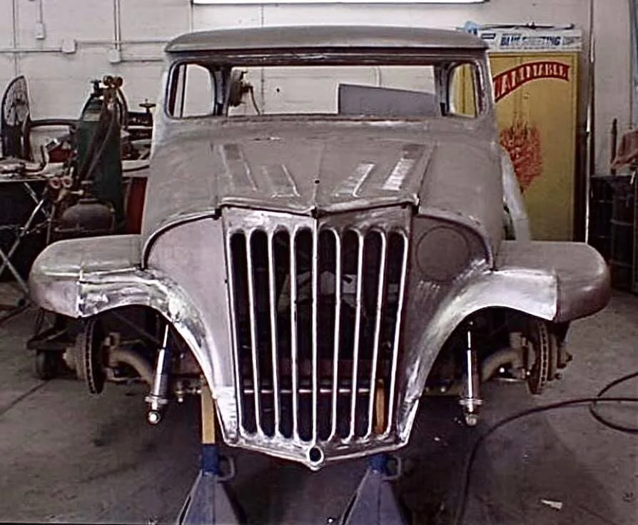 A vintage car body under restoration, with exposed metal and no paint, seen in a workshop with tools and equipment around.