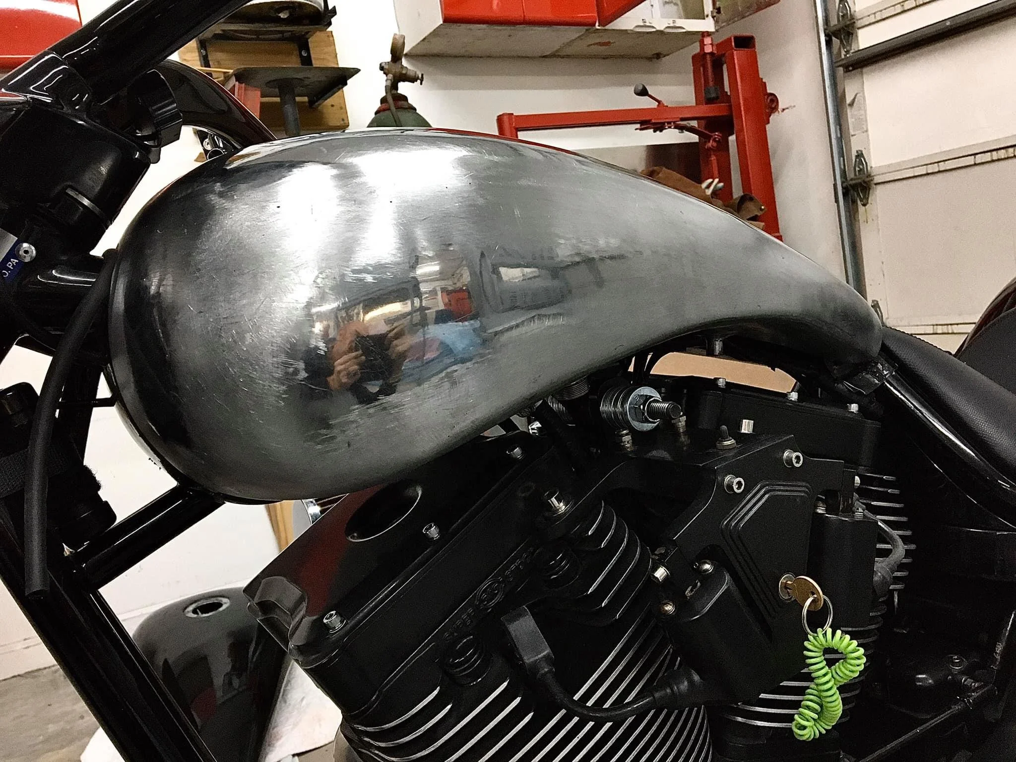 Close-up of a motorcycle's fuel tank and engine in a garage, with a reflection of the person taking the photo visible on the shiny metallic tank.