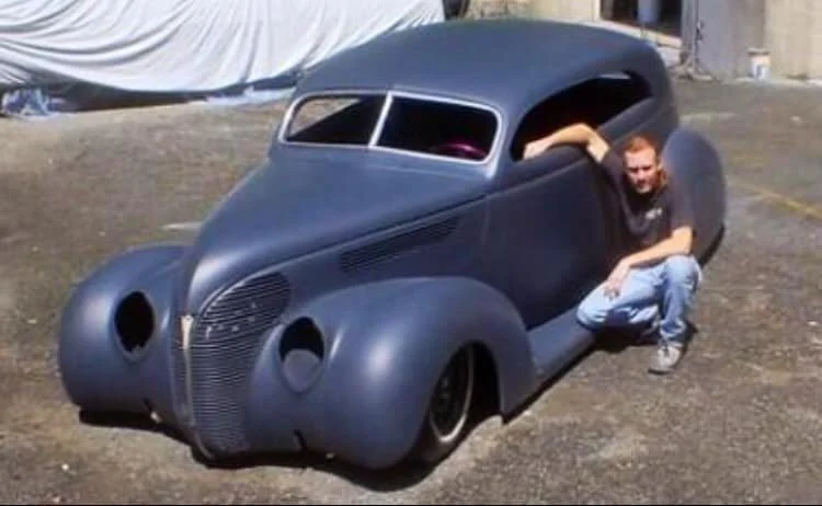 A man crouching next to a vintage black car with a unique, rounded design, parked on a gravel surface outside.