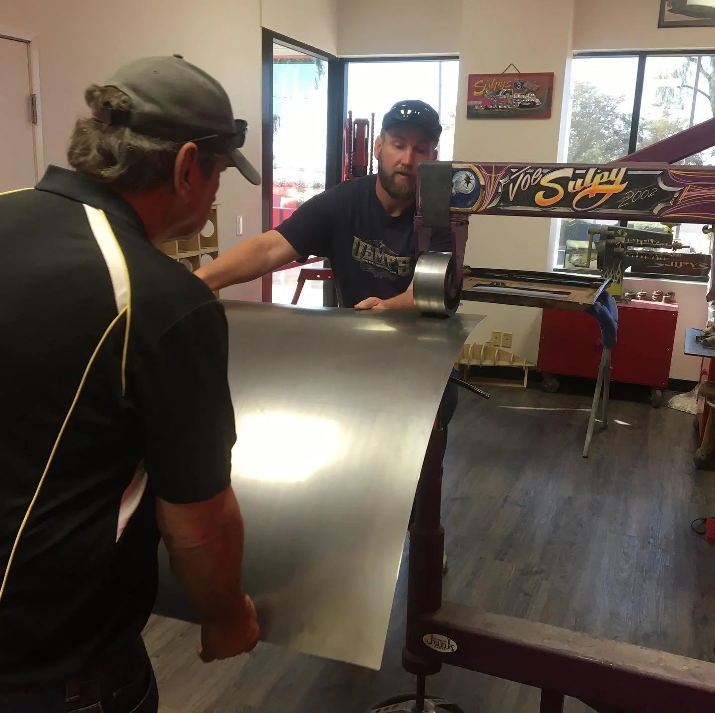Two men working together on a large metal sheet in a workshop, with one man holding the sheet and the other operating a machine.