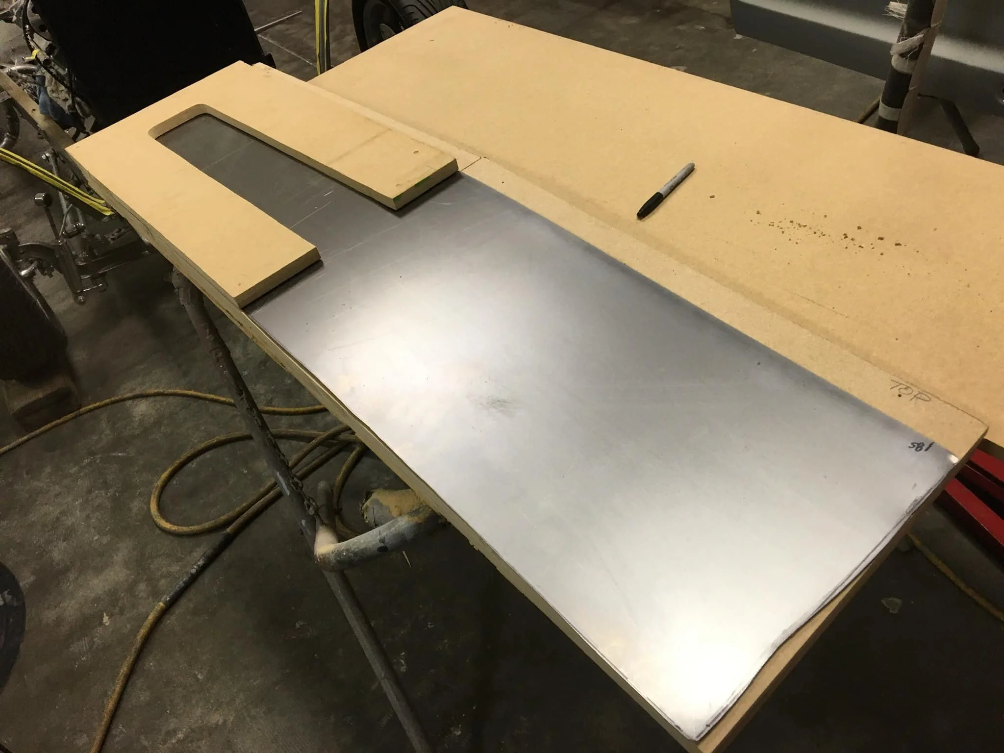 A work table with a large sheet of metal, pieces of wood, a marker, and some markings. The table is in a workshop or industrial setting.