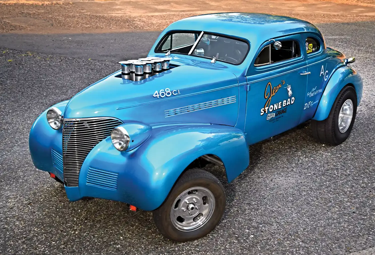 Blue vintage race car with racing decals and headers on the hood, parked on asphalt.