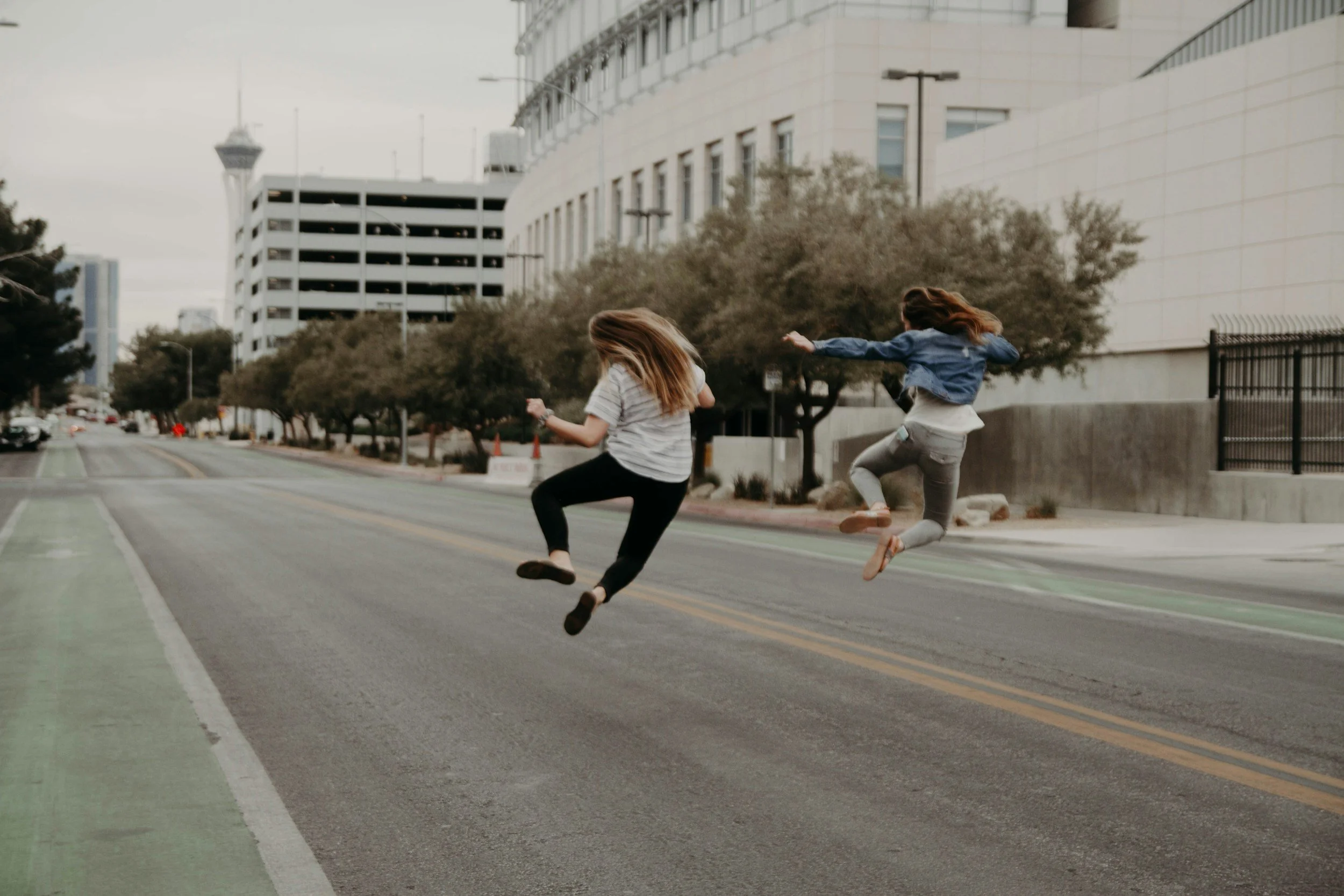 Two girls jumping in mid-air on city street, with modern buildings and trees in background.
