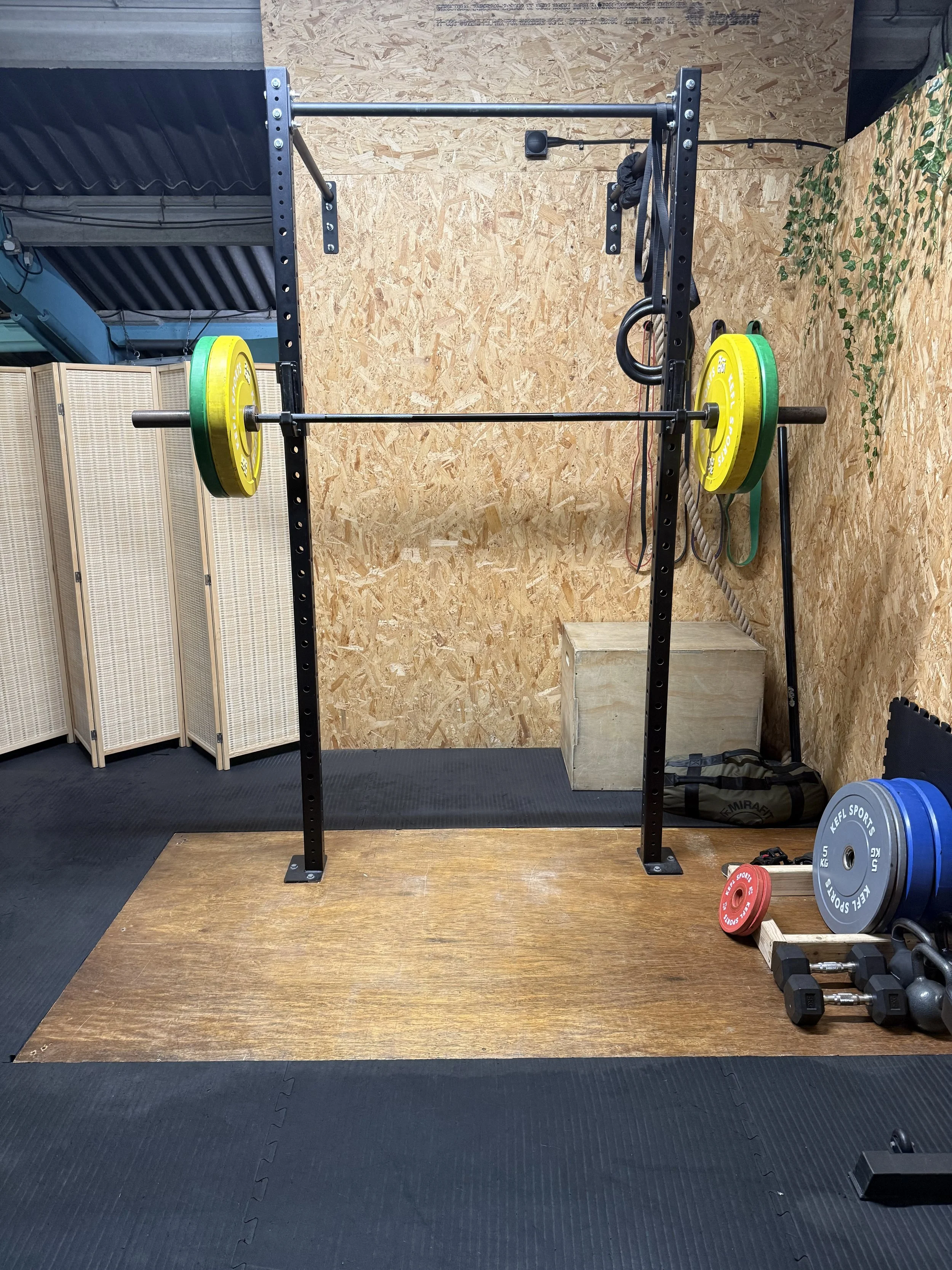 A weightlifting setup with a black pull-up bar rack, yellow and green weight plates on a barbell, and additional weights and gym accessories on the floor in a gym with wooden and black flooring.