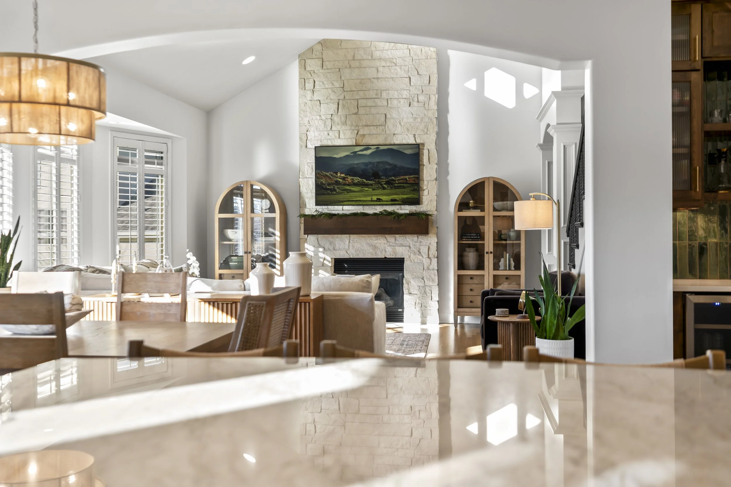 Bright living room with a stone fireplace, mounted TV, wooden cabinets, sofas, and large windows with white blinds.