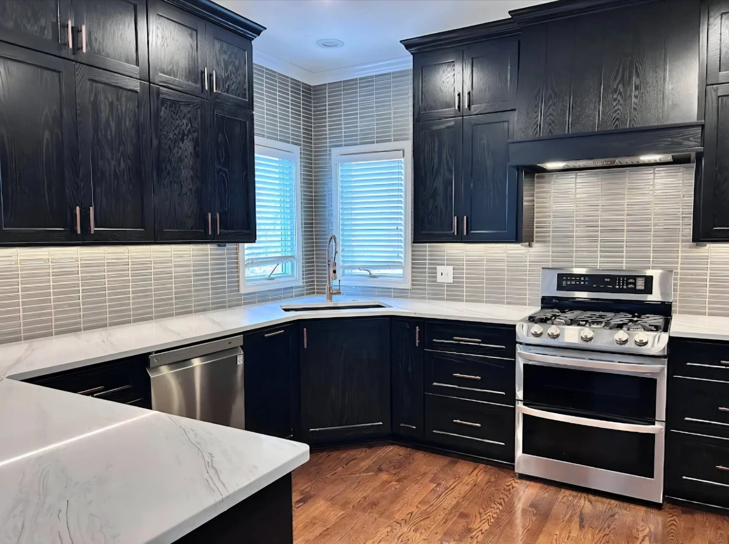 Modern kitchen with dark upper and lower cabinets, white stone countertops, stainless steel appliances, tiled backsplash, and two windows with white blinds.