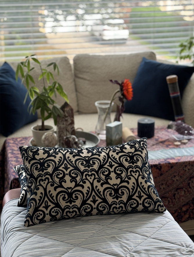 Interior view of a living room with a sofa, colorful pillows, a table with plants and decorative objects, and a large window with blinds.
