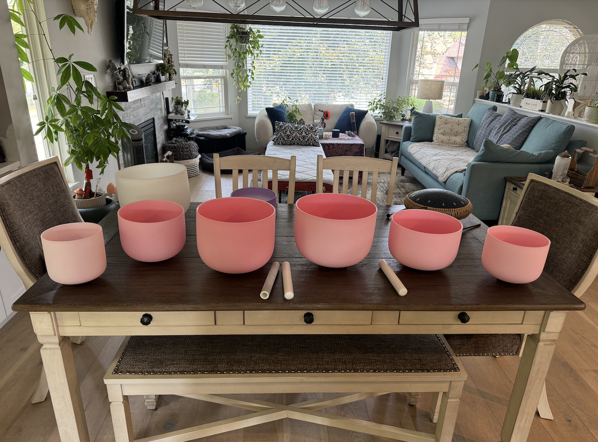 A wooden dining table with six pink and white singing bowls of various sizes and two mallets on top, in a bright living room with sofas, plants, and large windows.