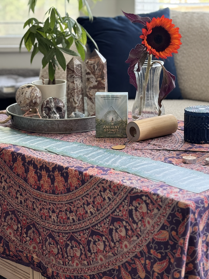 A table with a patterned tablecloth holds a potted plant, decorative skull stones, a tarot card deck, a crystal wheel, a rolled-up scroll, coins, a sunflower in a glass vase, and a blue container. A window with sunlight is in the background.