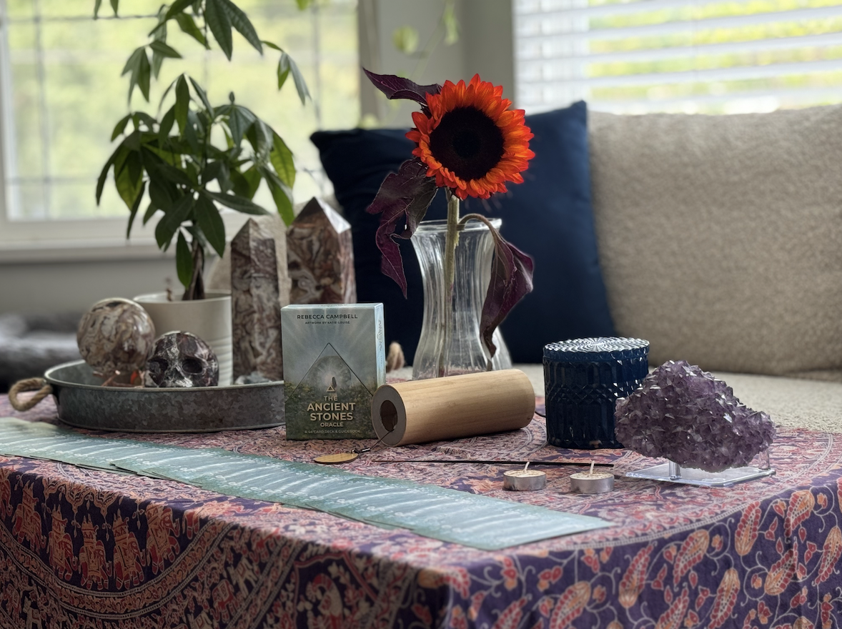 A table with a pink and purple patterned cloth, holding a purple amethyst crystal, a blue container, teal candles, and a deck of oracle cards titled 'The Ancient Stones' with an image of a triangle and a gemstone on the cover. Behind, there is a sunf