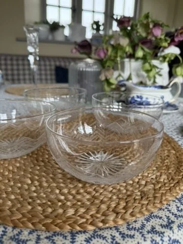 Clear glass bowls on a woven placemat on a table with a blue and white patterned tablecloth, with a floral arrangement and tableware in the background.