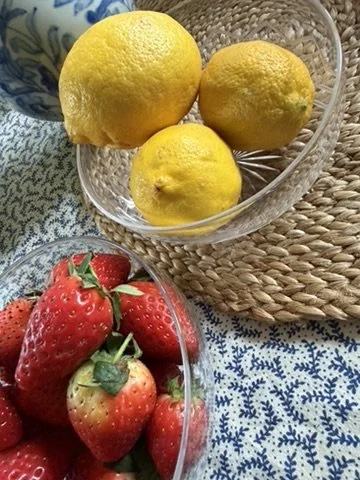 Three yellow lemons in a glass bowl on a woven placemat, with a pile of fresh strawberries in a clear bowl nearby on a blue and white patterned cloth.