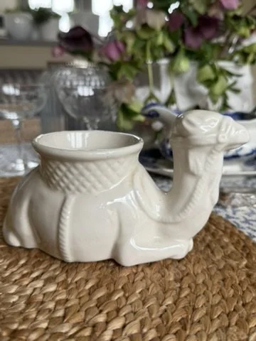 White ceramic camel-shaped container with a textured upper section, placed on a woven surface with flowers and glassware in the background.