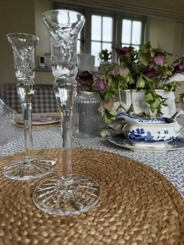 Close-up of two tall, empty glass candlesticks on a woven placemat on a dining table, with floral arrangements and dishes in the background.