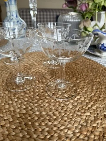 Three elegant glass dessert cups on a woven placemat, with a background of decorative vases, flowers, and a table setting.