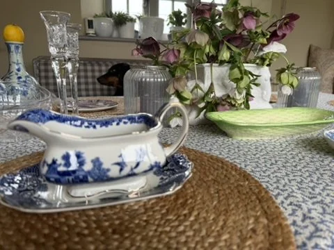 A dining table set with a blue and white porcelain gravy boat on a matching plate, placed on a woven placemat. There are pink flowers in a glass vase and various glassware in the background.