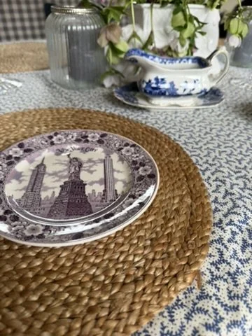Decorative table setting with a plate featuring the Statue of Liberty, placed on a woven placemat, with a blue and white gravy boat and floral arrangement in the background.