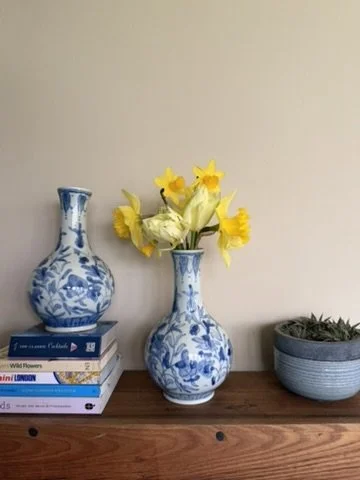 Blue and white floral vases with yellow and white flowers, a potted plant, and a stack of books on a wooden surface against a beige wall.