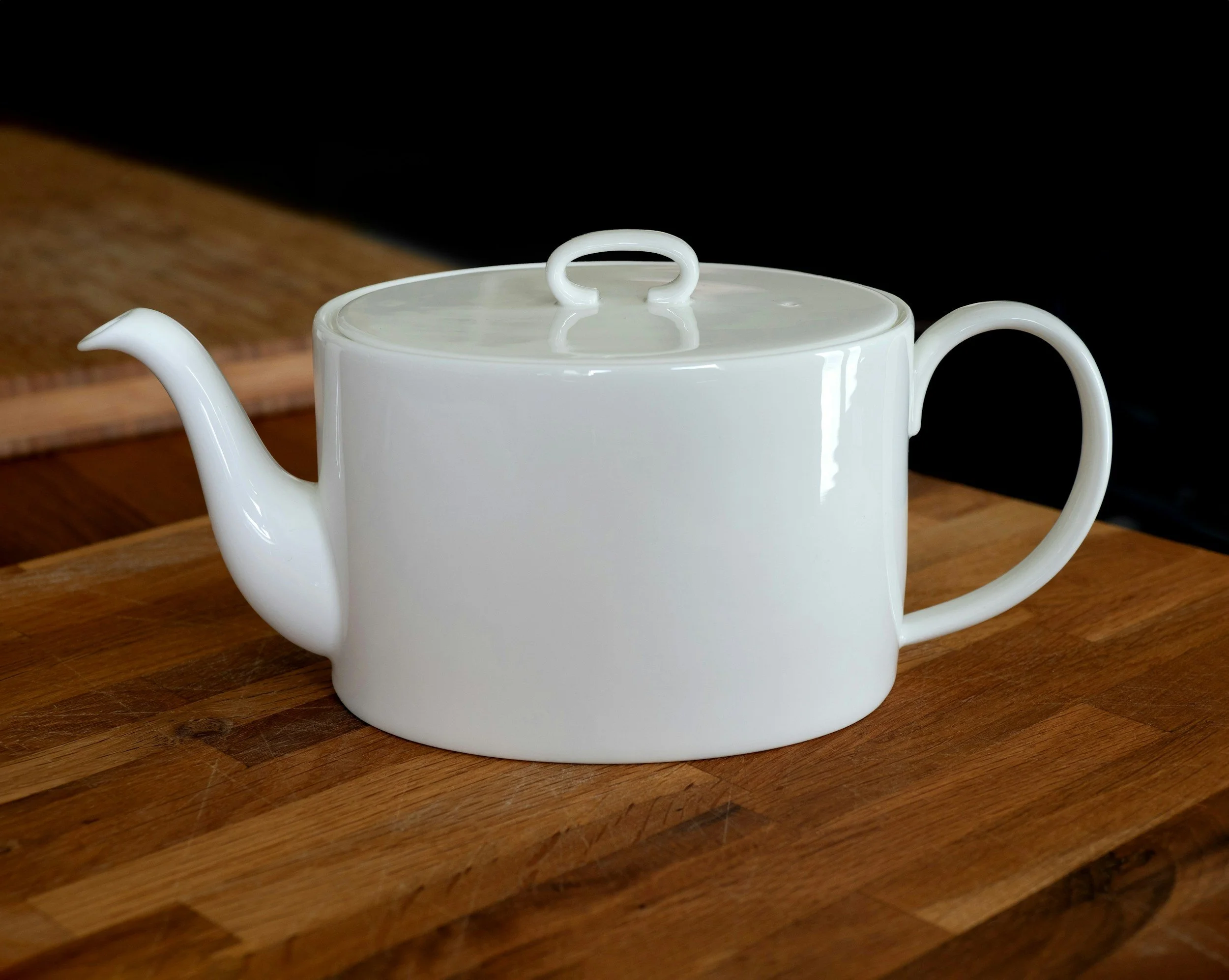 White ceramic teapot with a round handle on the lid, positioned on a wooden surface with a dark background.