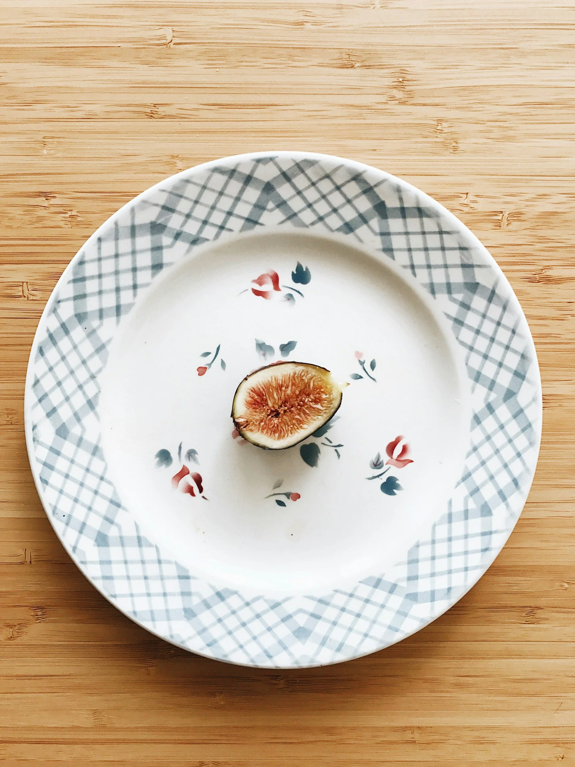 A plate with a floral pattern and a checkered border, holding a halved fig, on a wooden surface.