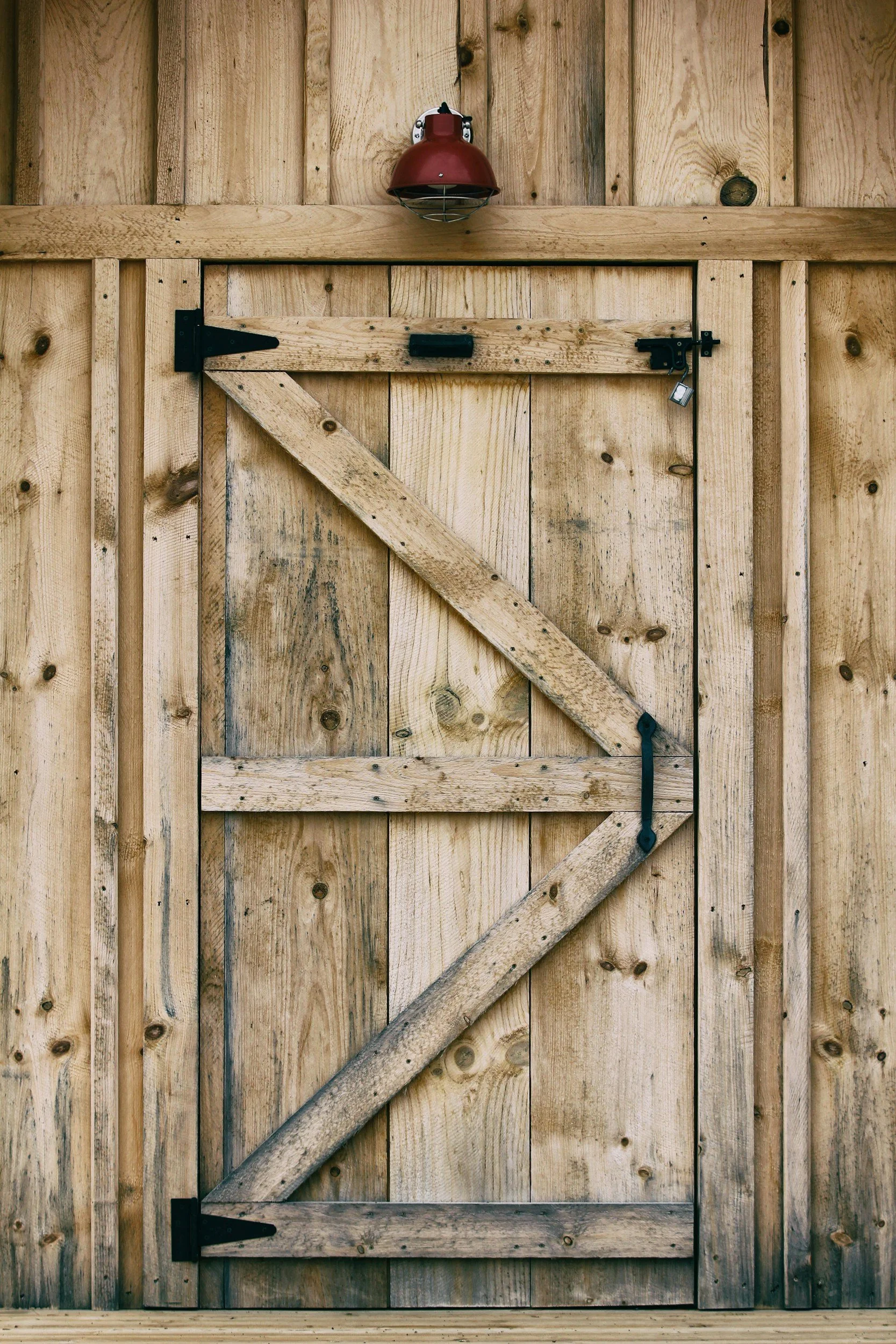 Wooden barn door with diagonal cross-brace, black metal hinges, and a red lantern-style light fixture mounted above.