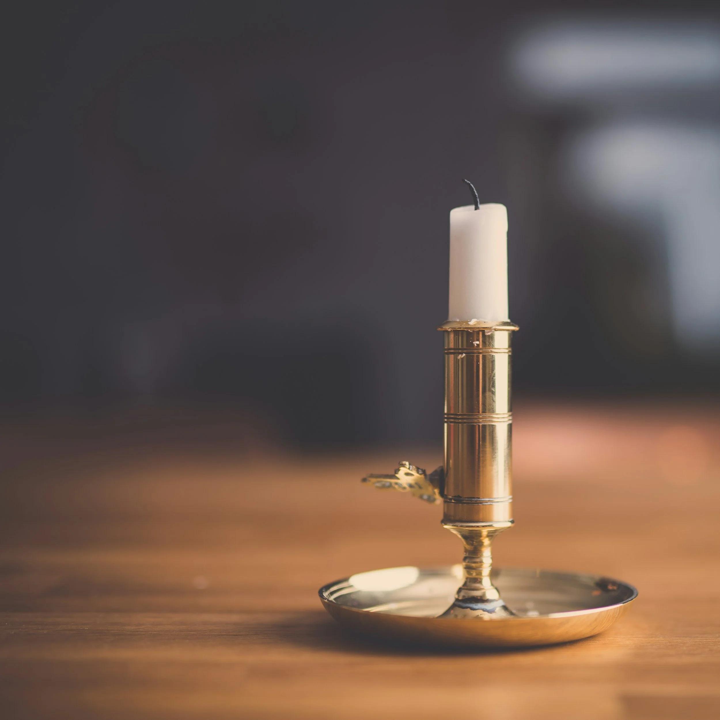 A close-up of a candle with a black wick, placed on a reflective metal dish, with blurred background and warm lighting.