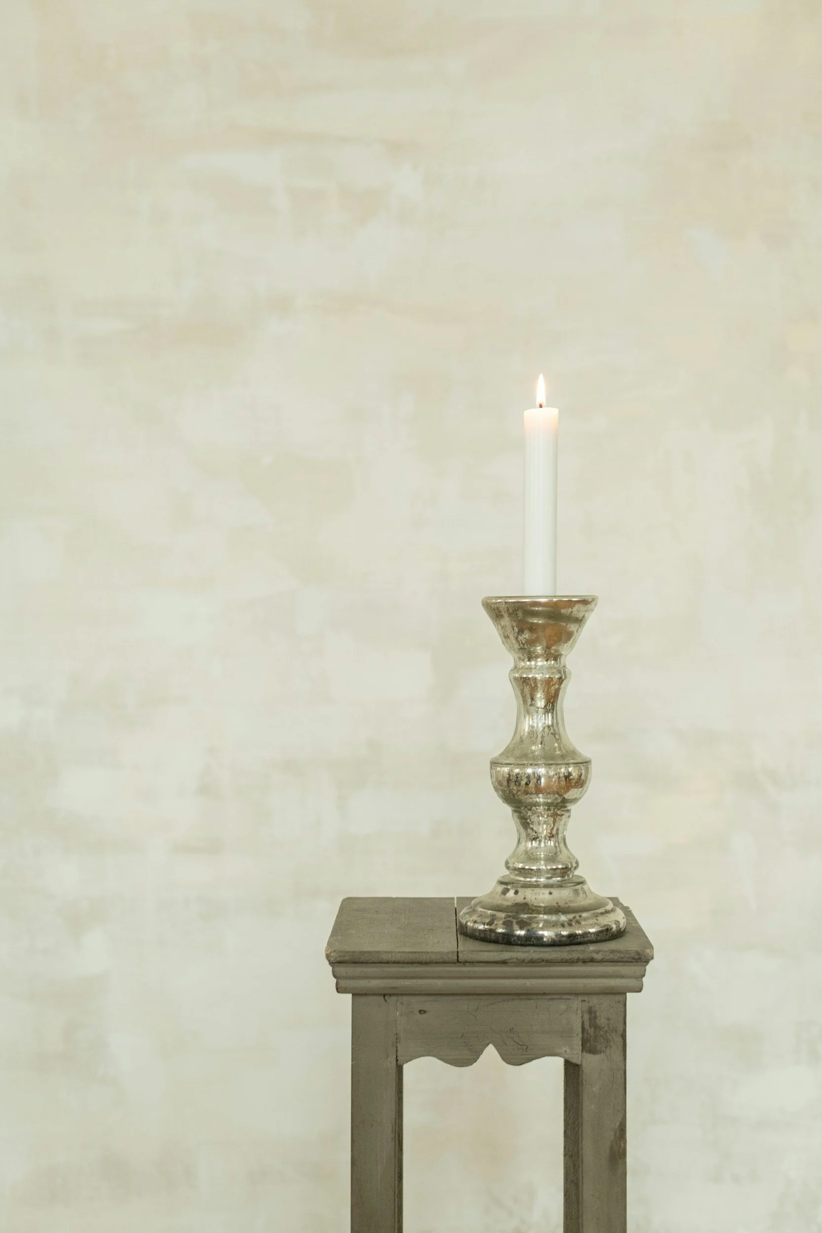 A lit white candle on a silver candle holder, placed on a gray wooden table in front of a pale yellow wall.