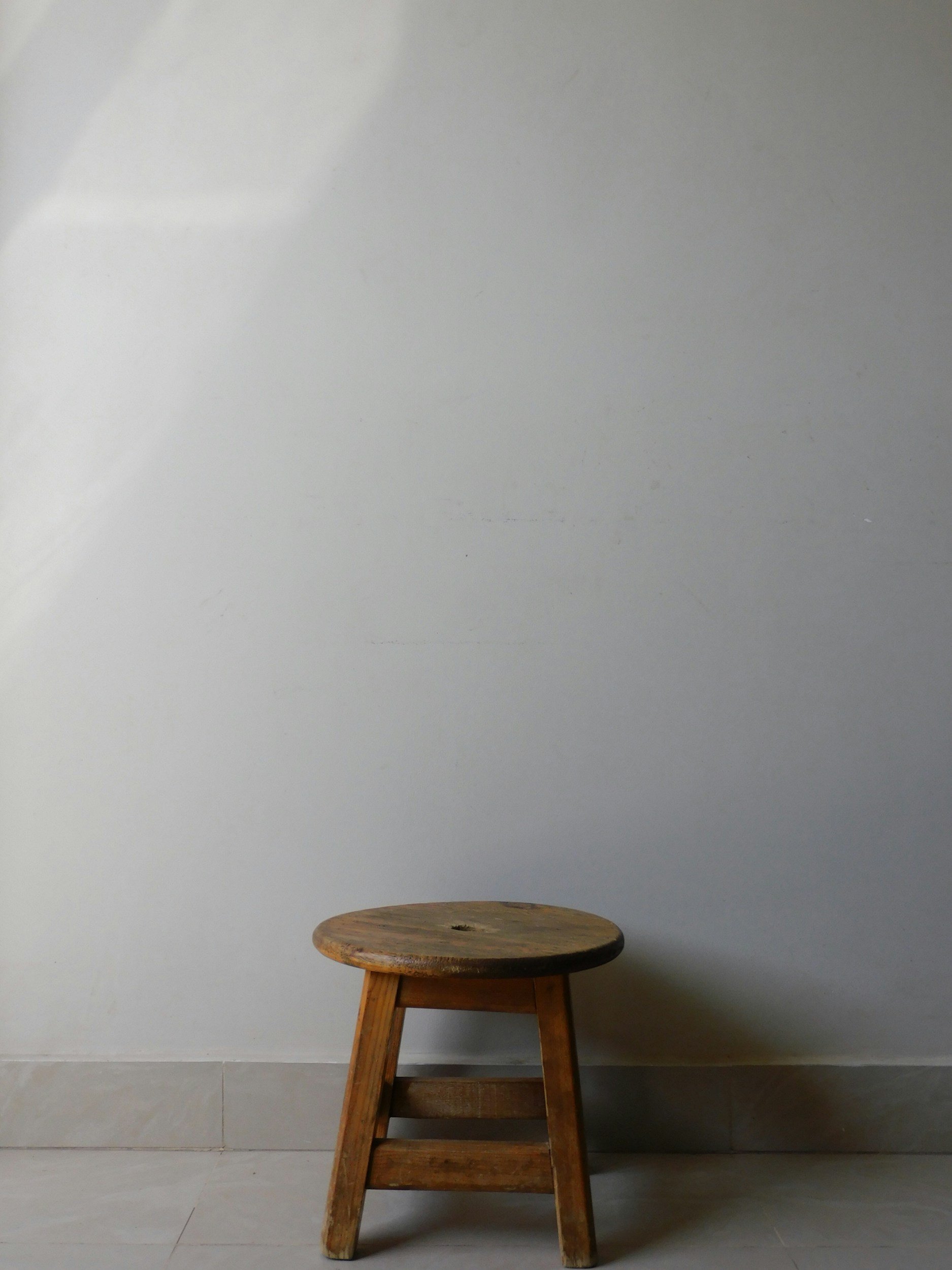 A wooden stool with three legs and a round top, placed against a plain wall.