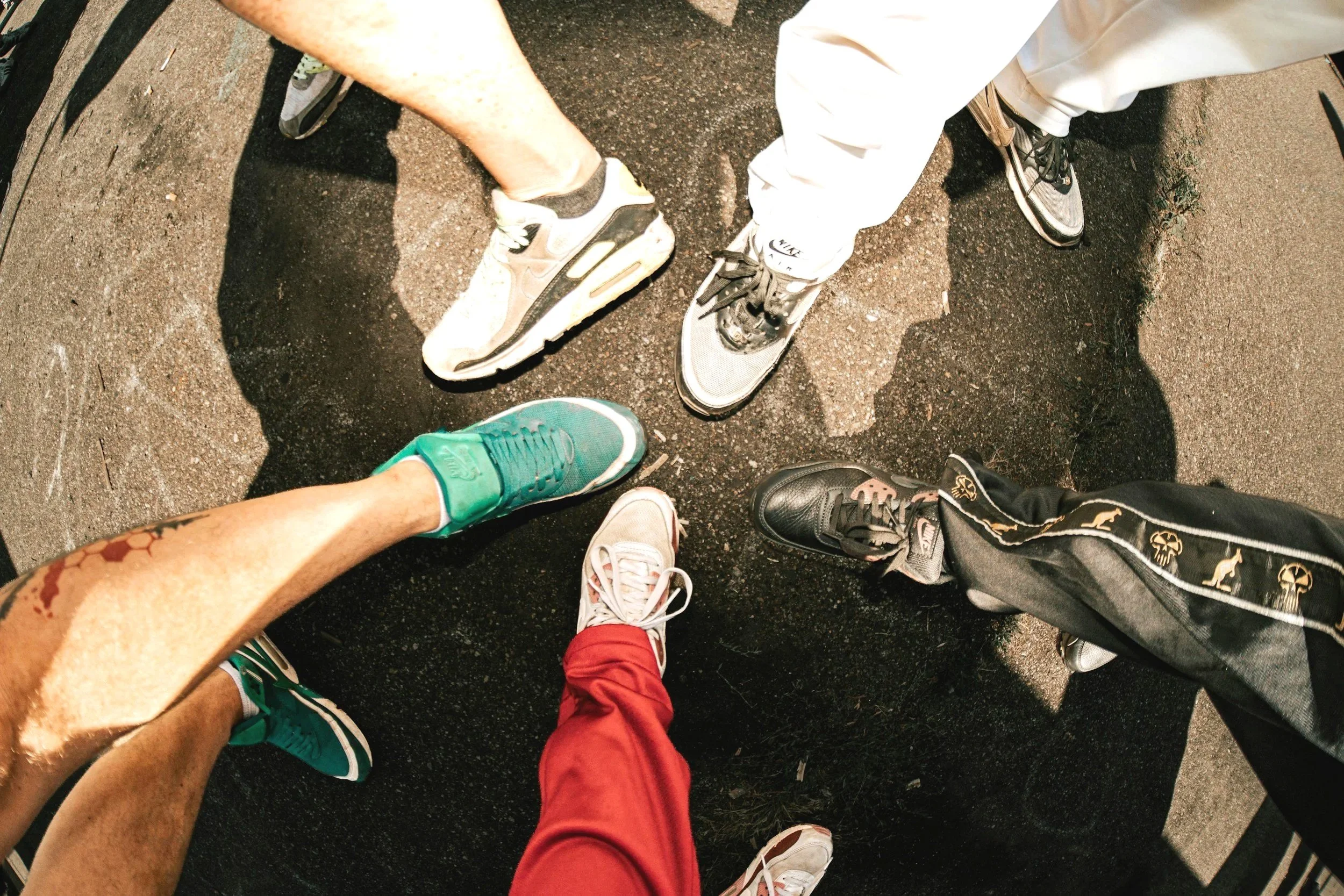 A top-down view of several people standing in a circle, showing their shoes and lower legs on a gravel ground.