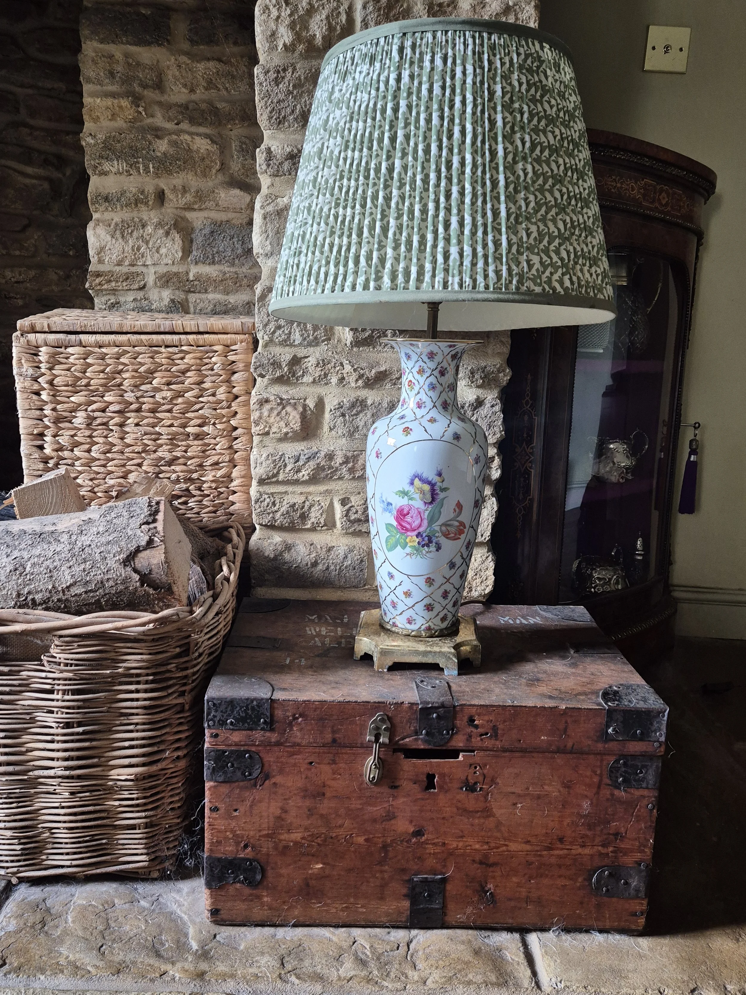 A decorative table lamp with a floral ceramic base and a pleated fabric lampshade with green and white patterns, placed on a weathered wooden chest next to stone walls and woven baskets.