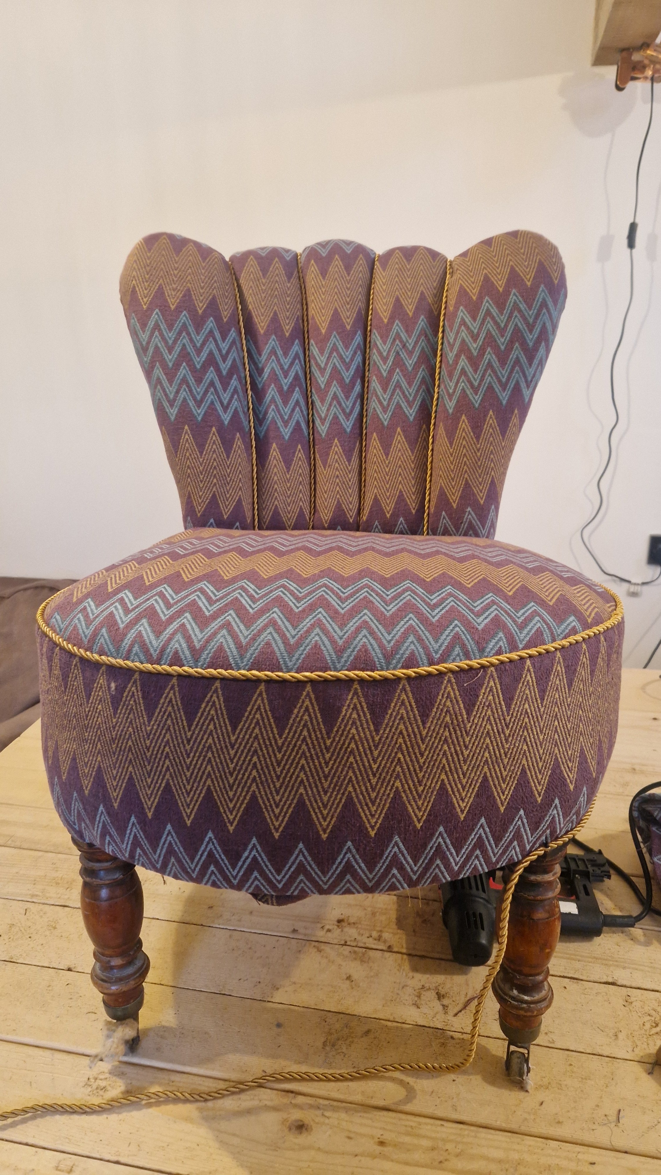 A vintage upholstered chair with a round, cushioned seat and a high, winged backrest, covered in fabric with a zigzag pattern in purple, blue, and gold, with wooden legs and a yellow trim around the edges.