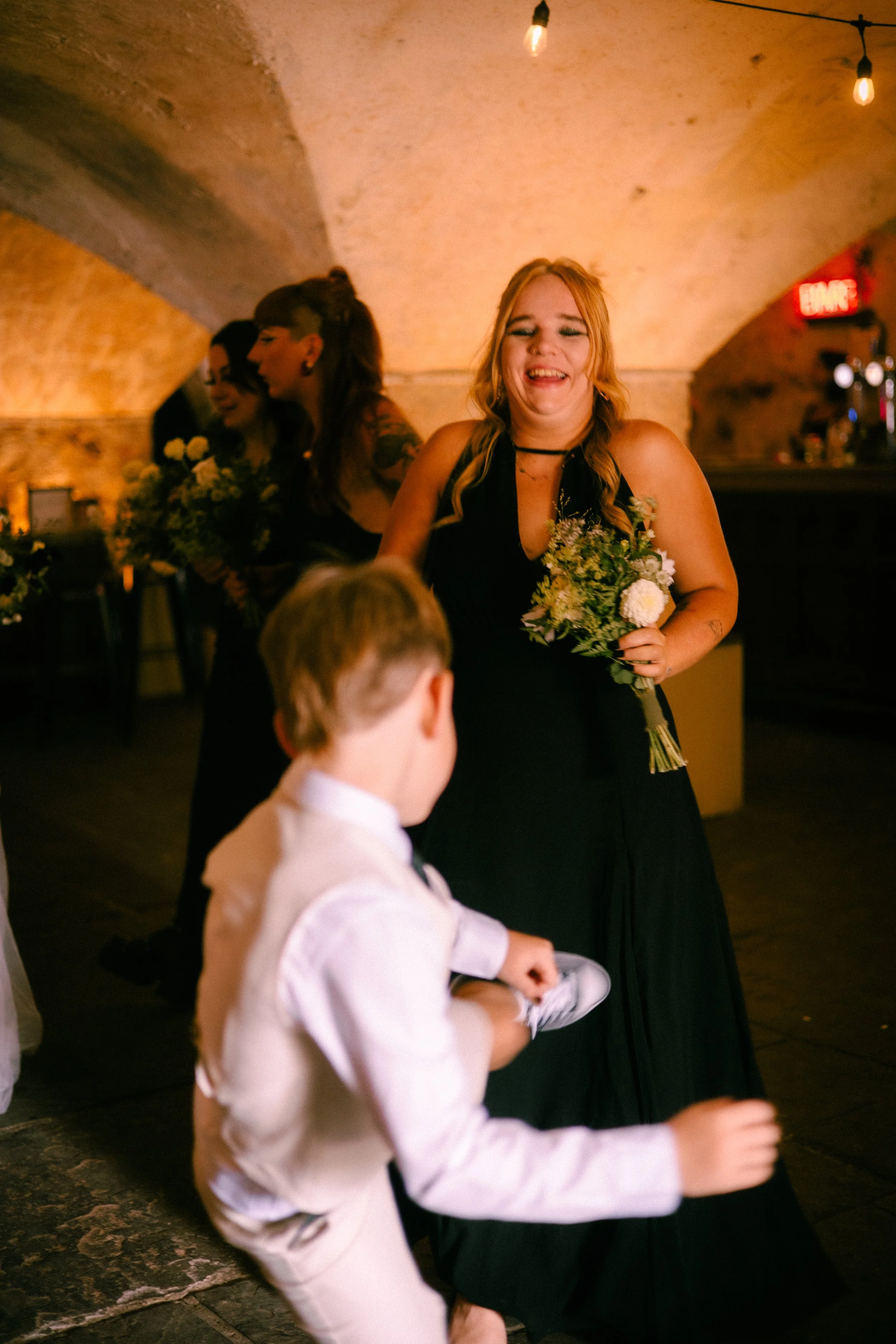 A woman with long blonde hair, smiling, holding a bouquet of flowers, wearing a black dress. In the foreground, a young boy wearing a white shirt with a vest is walking. In the background, other women and a bar area with an "EXIT" sign, hanging light