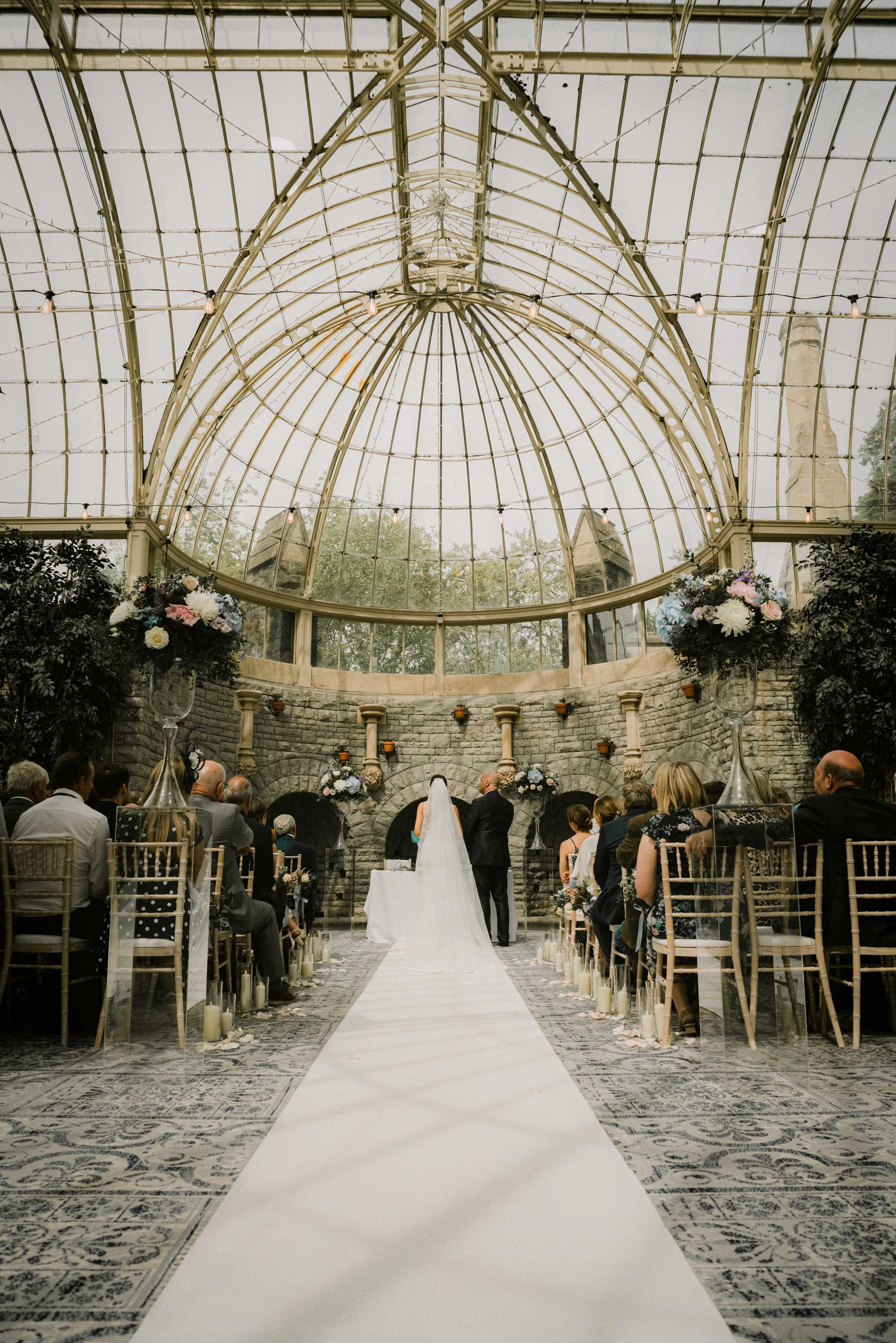 Indoor wedding ceremony with a bride and groom standing at the altar, surrounded by seated guests in a glass conservatory with ornate ceiling and stone wall backdrop.