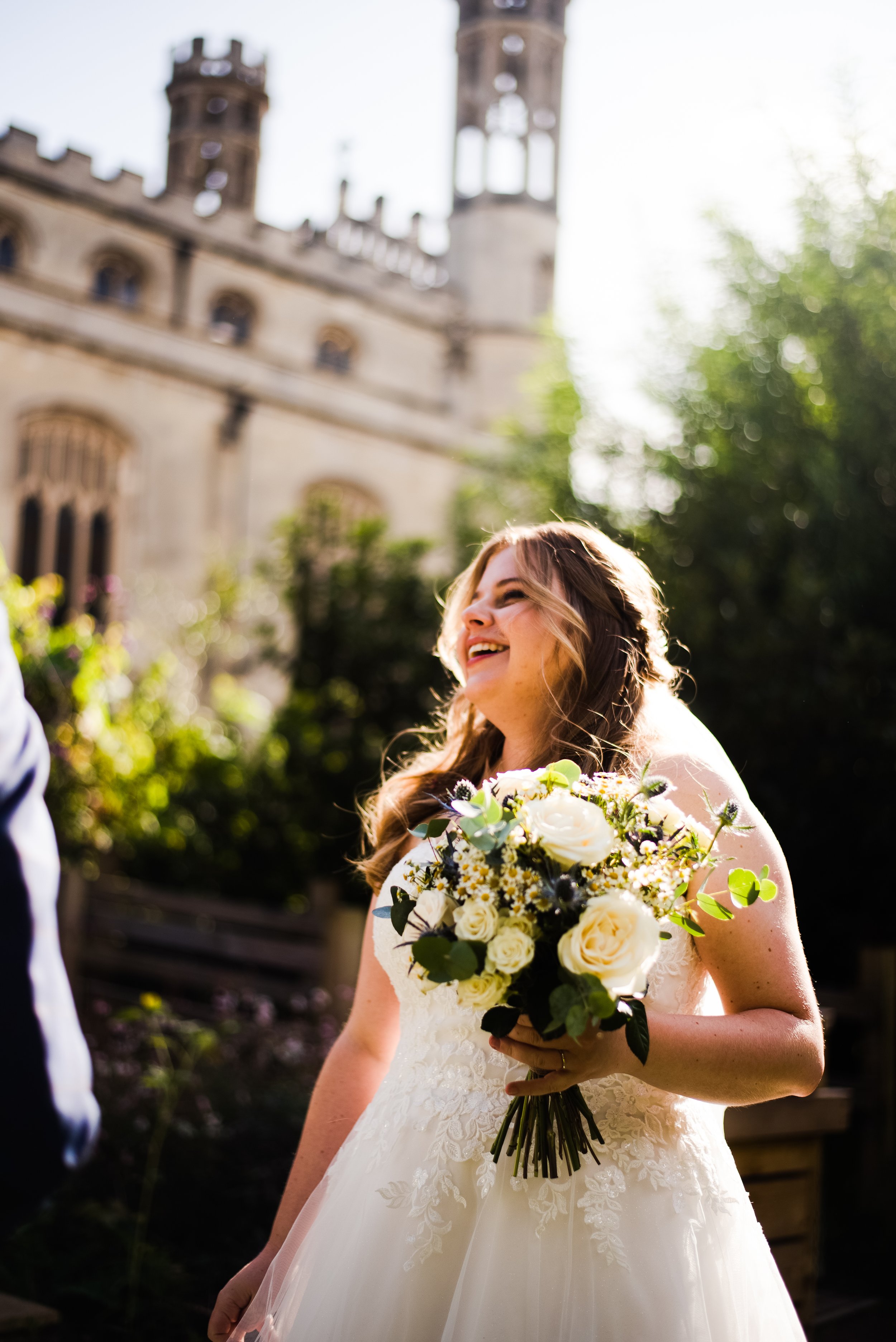 A smiling bride holding a bouquet of white roses and green foliage, standing outside near a historic castle with turrets, during daylight.