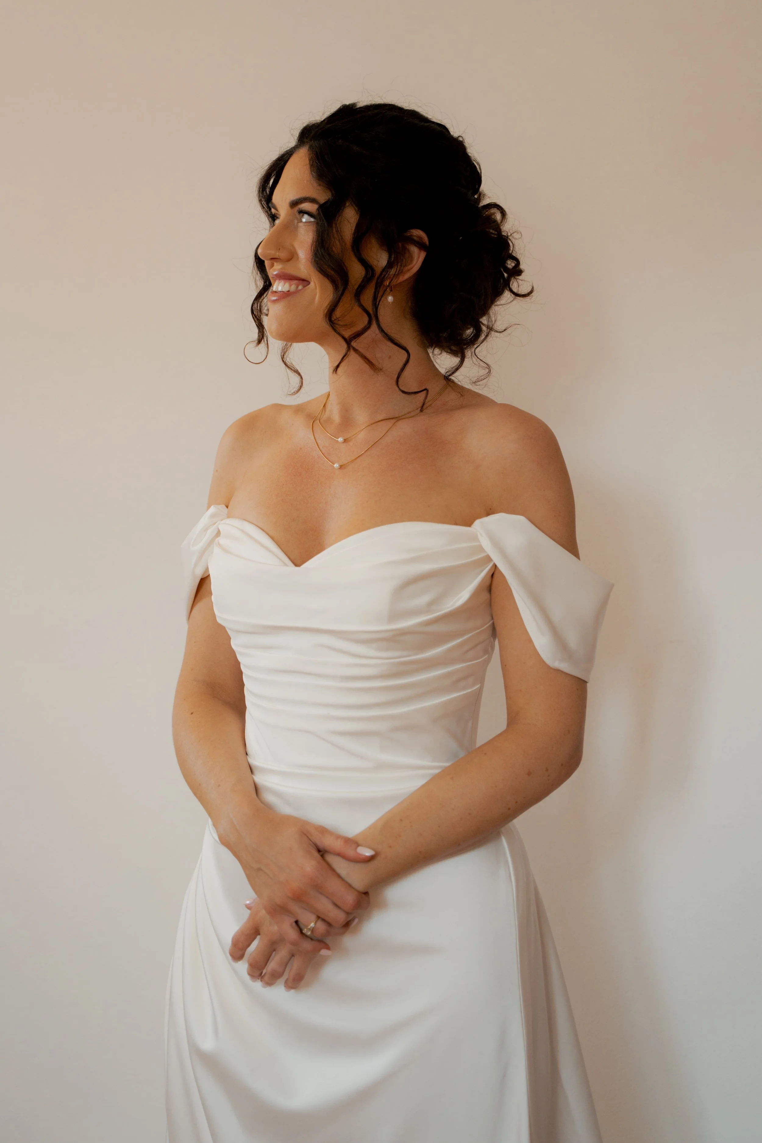 A woman in a white wedding dress with off-the-shoulder sleeves, standing against a plain beige wall, smiling and looking to her left.