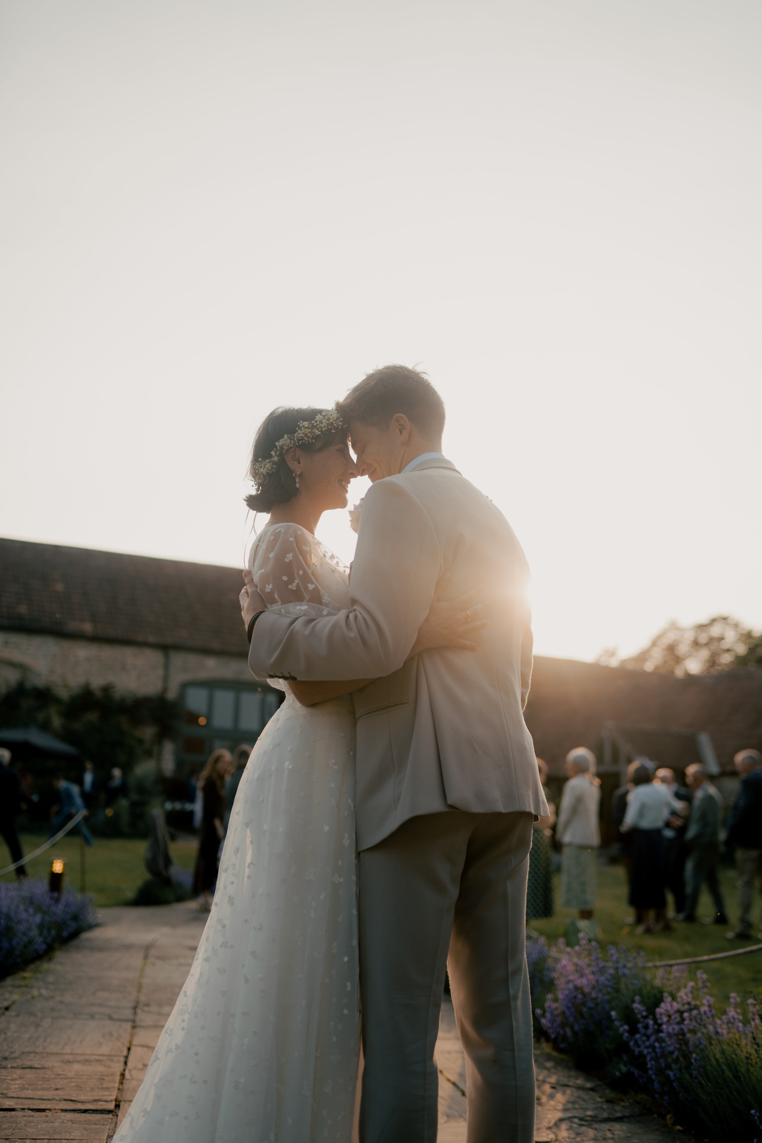A newlywed couple is sharing a kiss outdoors during sunset, surrounded by guests and lavender flowers.