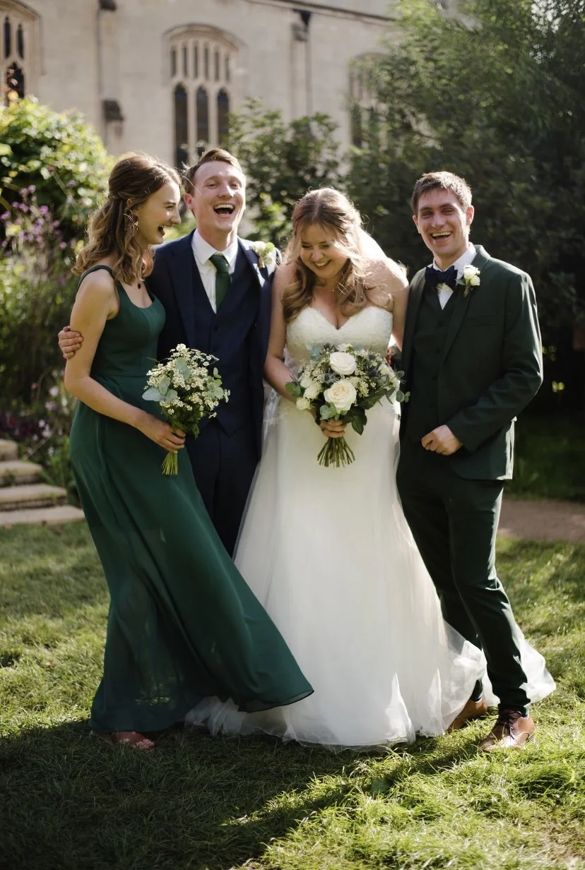 Group of five happy people at a wedding, with a bride in a white wedding dress holding a bouquet, and three friends, two men in suits and a woman in a green dress, standing on grass in front of a historic building.