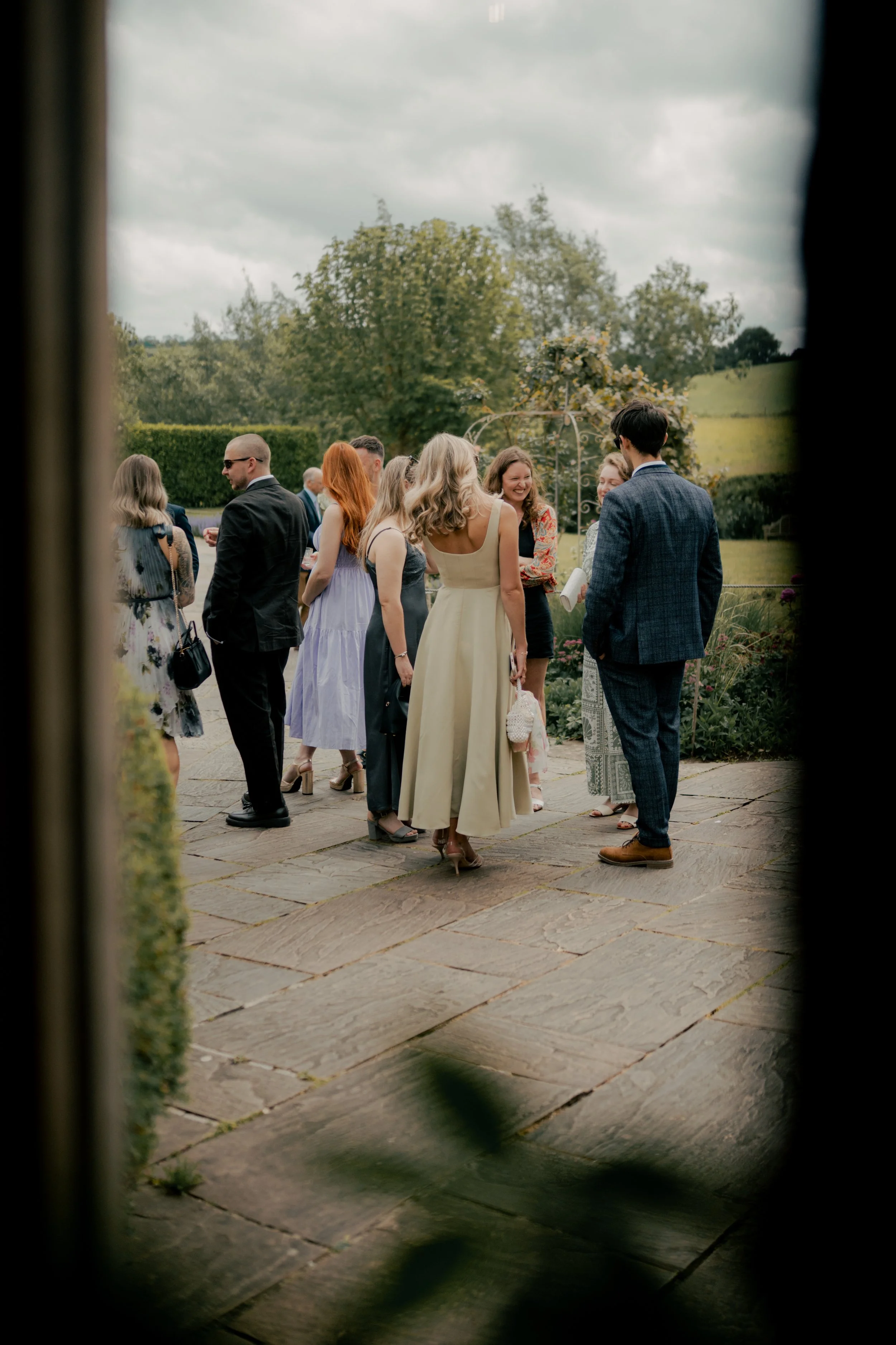 Group of people socializing outdoors at a garden party, seen through a window frame.