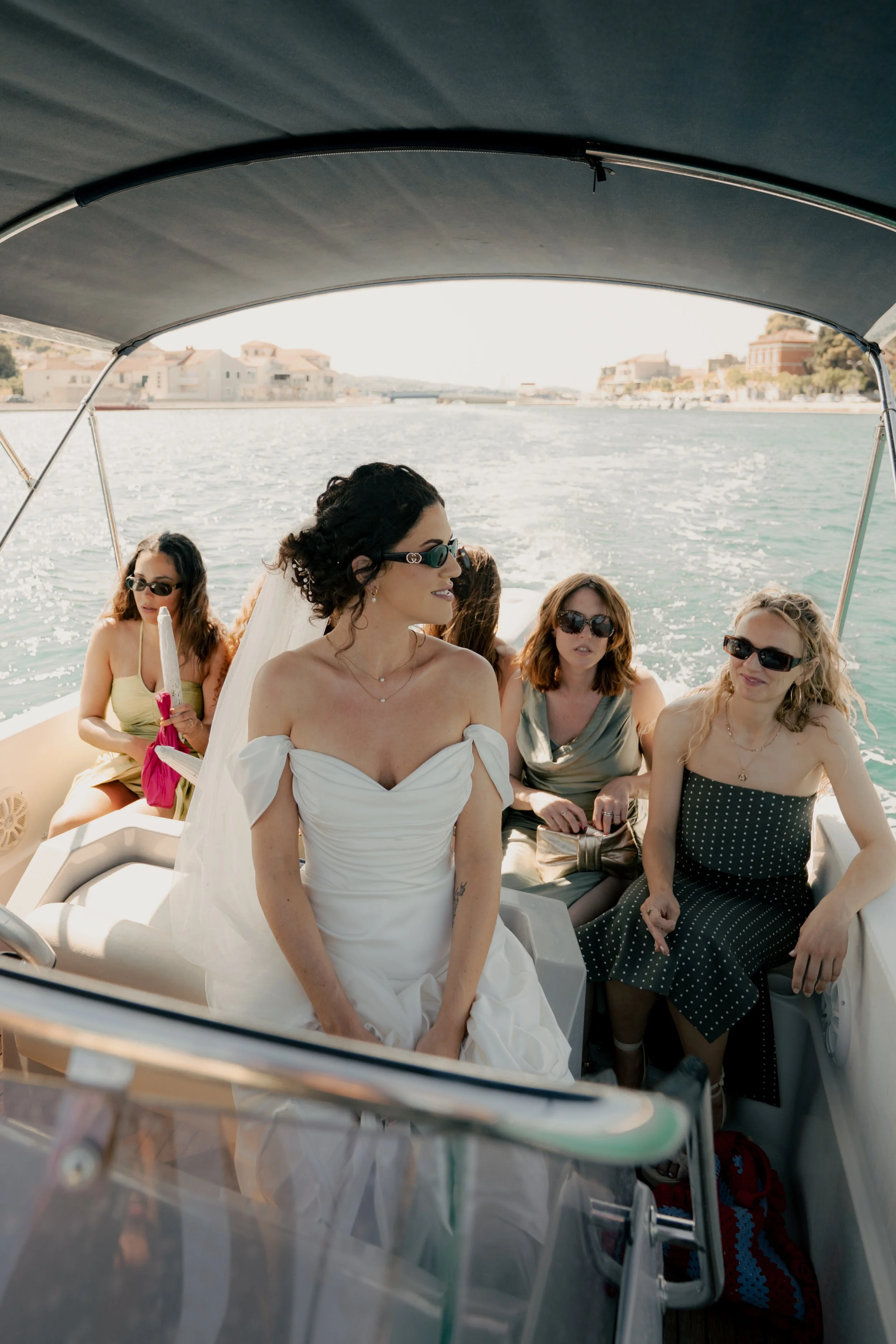 Women riding on a boat on the water, with a woman in a white dress and veil in the foreground, and other women sitting behind her, wearing sunglasses and summer dresses.