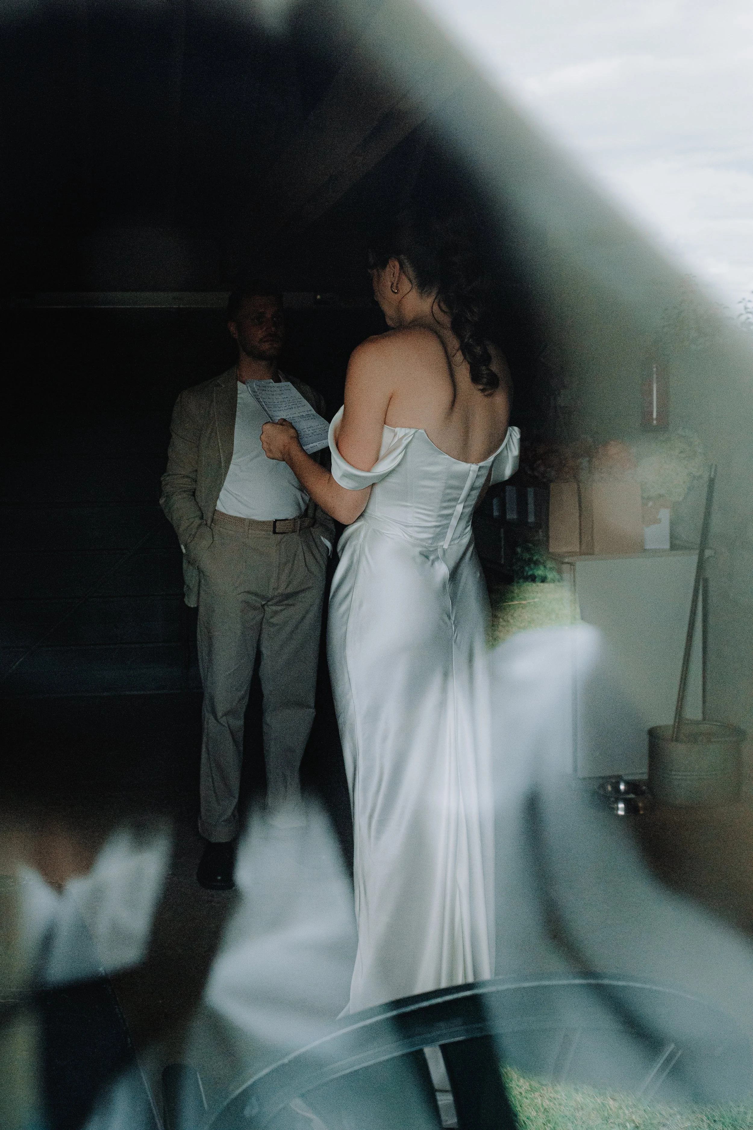 A woman in a white satin wedding gown with off-shoulder sleeves is reading a document while a man in a light-colored suit and white shirt stands beside her, both inside a vehicle.