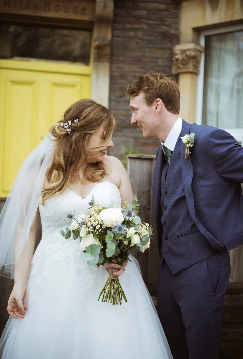 A bride and groom on their wedding day, facing each other and sharing a moment, with the bride holding a bouquet of white roses and greenery.